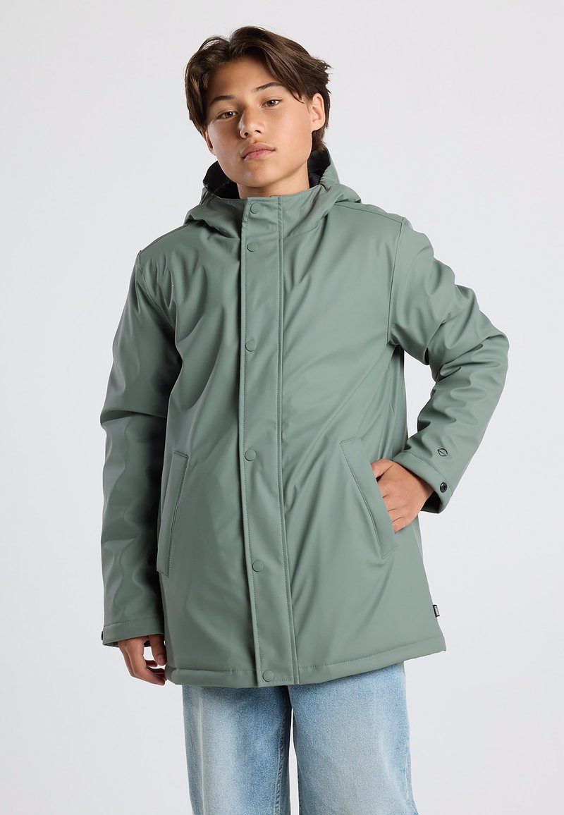 Green waterproof jacket with a hood, snap closures, and side pockets. Smooth texture and relaxed fit. Worn over light blue jeans.