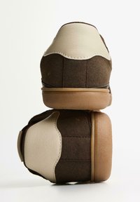 Une paire de baskets en daim marron avec un détail de talon en cuir beige et des semelles en caoutchouc caramel empilées verticalement, vues de dos.