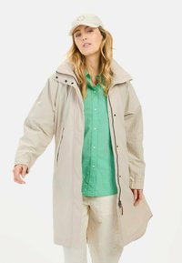 Helle beige lange Jacke mit hohem Kragen, Reißverschluss und Druckknöpfen, über einem grünen Hemd mit Knöpfen und hellen Hosen getragen.