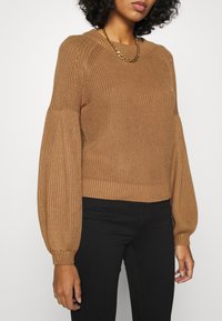 Brun stickad tröja med pösiga ärmar och ribbade manschetter, med rund halsringning och en något kortad passform. Högtalare med svarta byxor.