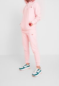 Persoon in een roze ellesse hoodie en bijpassende joggers met witte en teal sneakers, staande tegen een effen witte achtergrond.