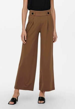 Vrouw die bruine wijde broek met hoge taille draagt met zwarte knoopdetails, gecombineerd met zwarte open teen mules met hak en een zwart mouwloos topje.