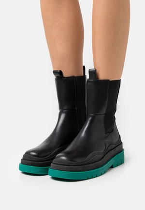 Botas de cuero negras hasta el tobillo con una parte superior texturizada, paneles laterales elásticos y una suela gruesa en color teal con patrones de relieve pronunciados.
