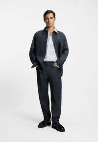 Dunkle Jeansjacke über einem weiß-navy gestreiften Shirt, kombiniert mit dunklen Jeans mit geradem Bein und schwarzen Schuhen. Sauberes, modernes Design.