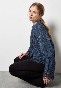 Camisa azul de botones hecha de tejido texturizado con intrincados adornos de encaje en los puños y el dobladillo, combinada con pantalones negros ajustados.