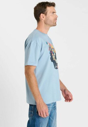 Mann im hellblauen T-Shirt mit farbenfrohem, floralem Totenkopf-Design, steht im Profil, trägt blaue Jeans vor weißem Hintergrund.