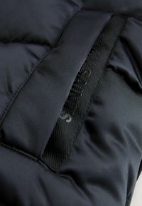 Svart vadderad jacka i tyg med en slät yta, med ett glänsande svart logotag integrerat i en kontrasterande vävd panel.