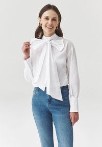 Blusa blanca de manga larga con un gran lazo en el cuello; tejido suave; combinada con jeans azules, mostrando una silueta ajustada.