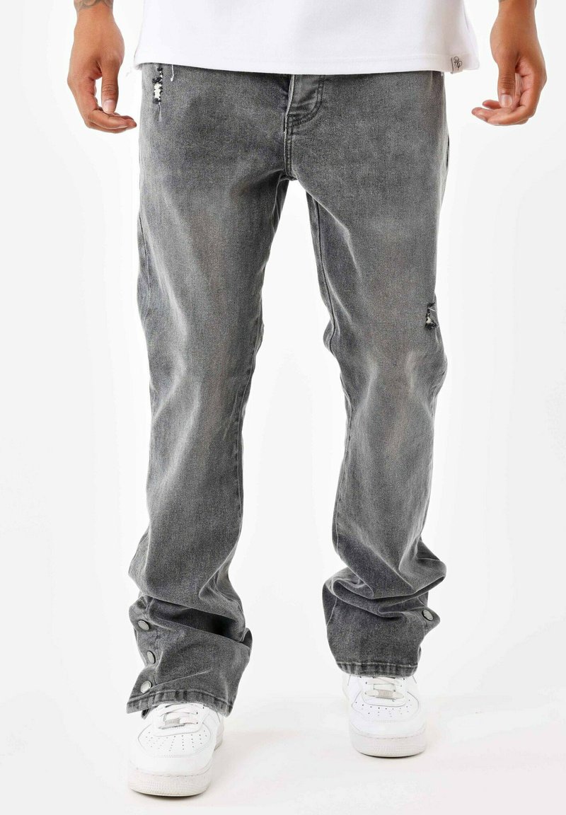 Jeans de mezclilla gris con un corte relajado, que presentan detalles desgastados, cierres a presión en los tobillos y una textura usada. Combinados con zapatillas blancas.