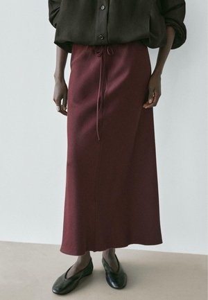 Persona con una camicia marrone scuro abbottonata, una gonna color borgogna alla caviglia con cintura in vita, scarpe nere piatte e un anello d'argento sulla mano sinistra.