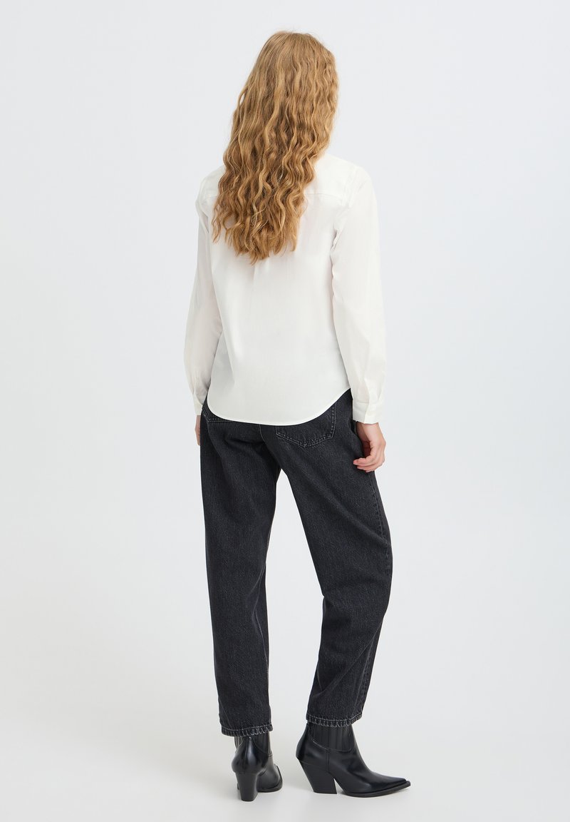 Femme aux longs cheveux ondulés portant une chemise blanche à manches longues, un jean noir ample et des bottines noires à talons, debout de dos.