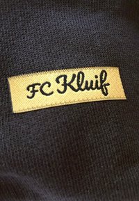 Zwarte stoffen patch met "FC Kluif" in zwarte cursieve tekst op een gouden achtergrond, in de stof gestikt.