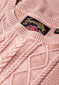 Pinker gestrickter Pullover mit Zopfmuster und einem gerippten Kragen. Das Etikett zeigt "SuperDry" in Gold mit einem Rosendesign.