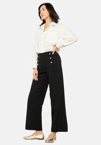 Črne hlače širokega kroja z zlatimi gumbnimi detajli, v kombinaciji z belo srajco in metaliziranimi zlatimi balerinkami. Gladka tekstura tkanine.