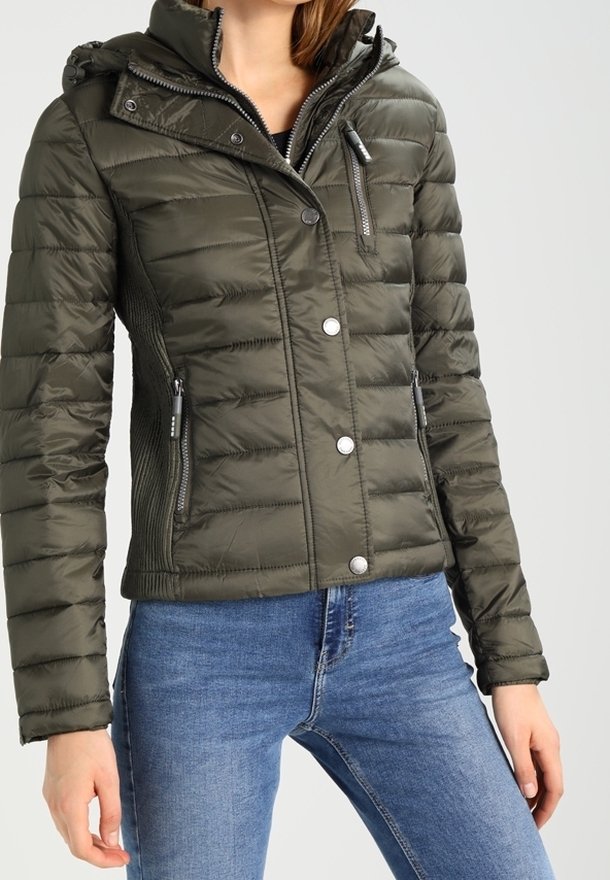 Femme portant une veste matelassée vert foncé avec fermeture éclair et boutons, associée à un jean bleu sur un fond uni.