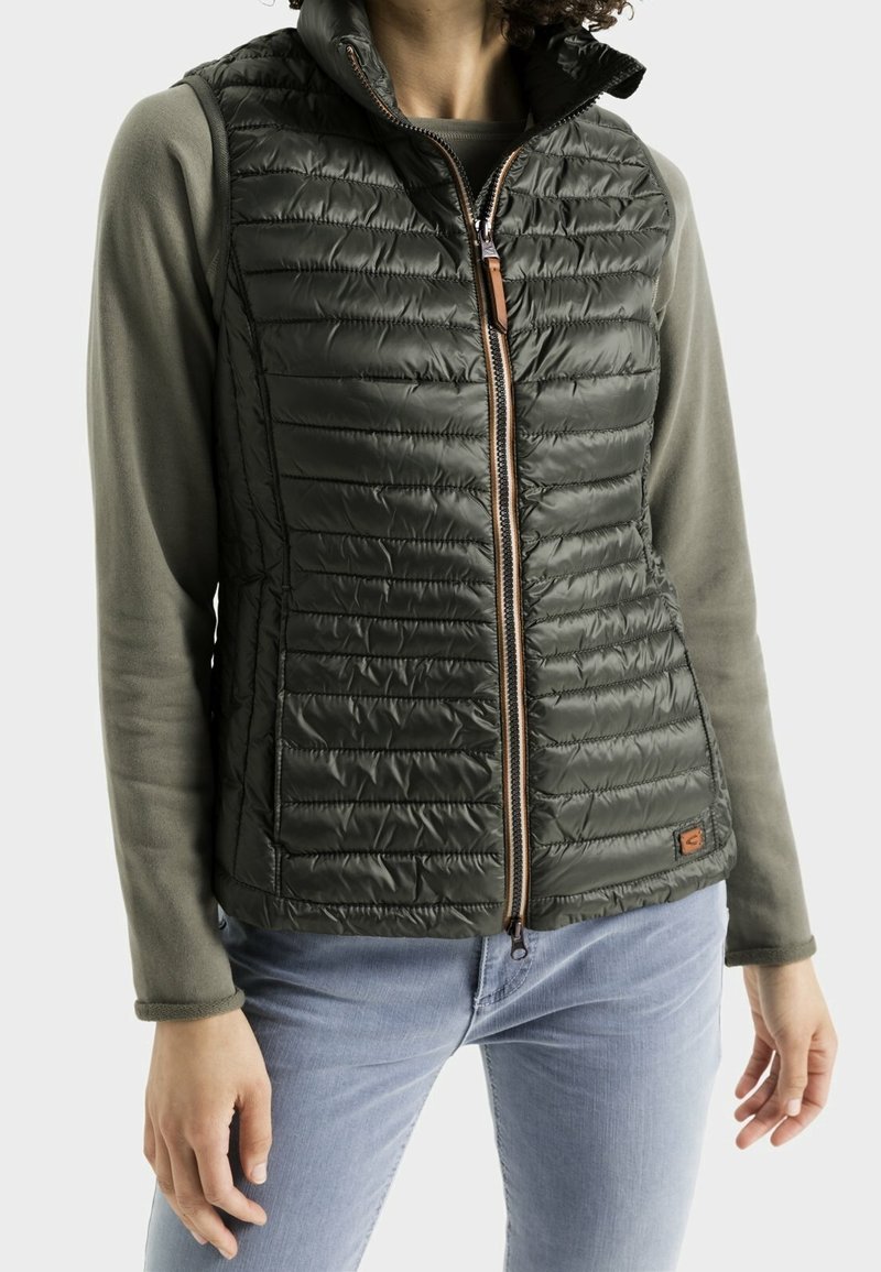 Gequiltede donkergroene vest met hoge kraag, ritssluiting aan de voorkant en bruin leren trekker. Draagt over een grijs overhemd met lange mouwen en blauwe jeans.