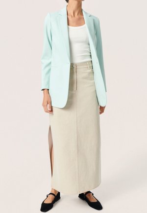 Vrouw draagt een lichtblauwe blazer, witte mouwloze top, beige lange rok met zijsplit en zwarte platte schoenen op een effen achtergrond.