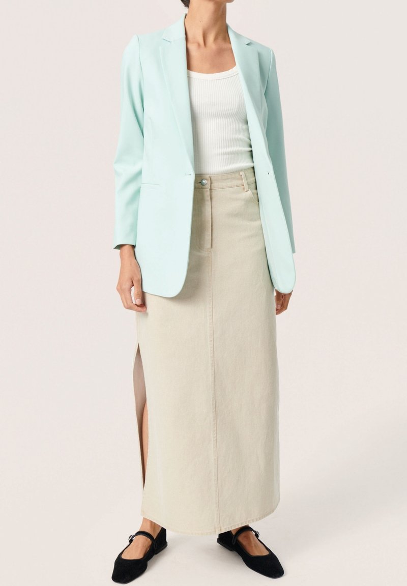 Femme portant un blazer bleu clair, un haut sans manches blanc, une longue jupe beige avec une fente latérale et des chaussures plates noires sur un fond uni.