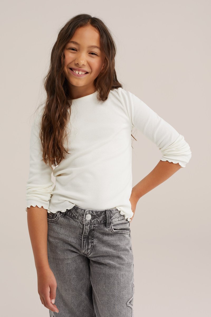 Lächelndes Mädchen mit langen braunen Haaren trägt ein weißes Langarmshirt und graue Jeans, posiert mit einer Hand auf der Hüfte vor einem einfarbigen Hintergrund.