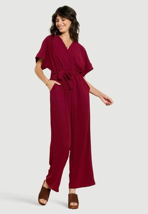 Vrouw draagt een bordeauxrode korte mouw jumpsuit met wijde pijpen en een tailleceintuur, poseert met één hand in de zak en bruine open-toe schoenen.