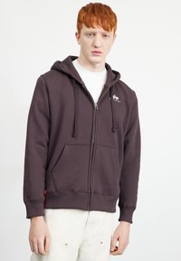 Sudadera con cremallera de color gris oscuro, hecha de una mezcla de algodón. Cuenta con bolsillos tipo canguro, puños acanalados y un pequeño logotipo en el pecho izquierdo.