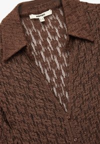 Brun blondelace cardigan med krave, der har et struktureret, åbent strikket mønster og fire knaplukninger foran.