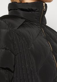 Czarna kurtka puchowa z wysokim kołnierzem, zapięciem na zamek i pikowanym wzorem. Posiada złote akcesoria i ukośne detale przeszycia.
