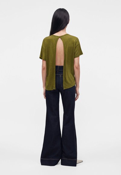 Personne aux longs cheveux lisses portant un top vert olive à manches courtes avec le dos ouvert et un jean foncé taille haute évasé, debout de dos.