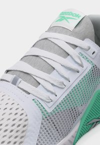 Κοντινό πλάνο λευκού αθλητικού παπουτσιού Reebok με πράσινες λεπτομέρειες, επάνω μέρος από αεριζόμενο δίχτυ και λευκά κορδόνια δεμένα πάνω από τη γκρι γλώσσα.