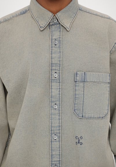 Ljusblå denimskjorta med knäppning fram, spetsig krage och en bröstficka. Har sömnadsdetaljer och en subtil tygtextur.