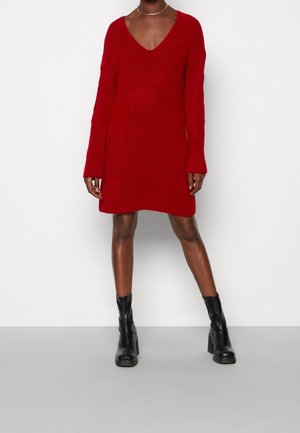 Vestito maglione rosso lavorato a maglia con scollatura a V profonda e maniche lunghe, caratterizzato da una vestibilità comoda e polsini a costine. Indossato con stivali alla caviglia neri con il tacco.