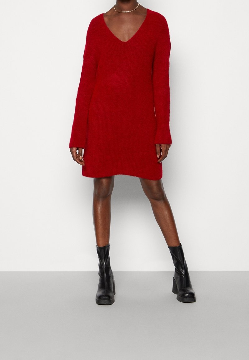 Robe pull rouge tricotée avec un décolleté en V profond et des manches longues, présentant une coupe décontractée et des poignets côtelés. Portée avec des bottines à talons noires.