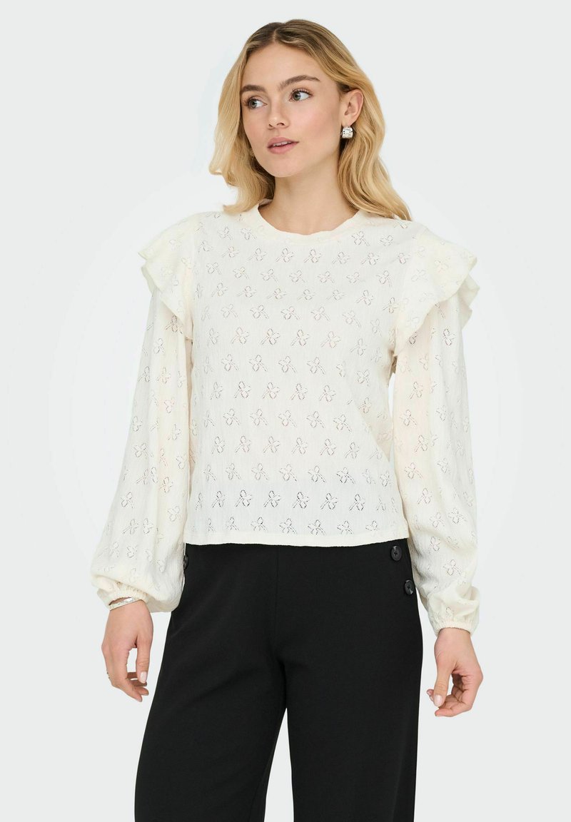 Jeune femme portant un chemisier en dentelle blanche avec des épaules volantées et un pantalon noir taille haute, regardant sur le côté sur un fond uni.