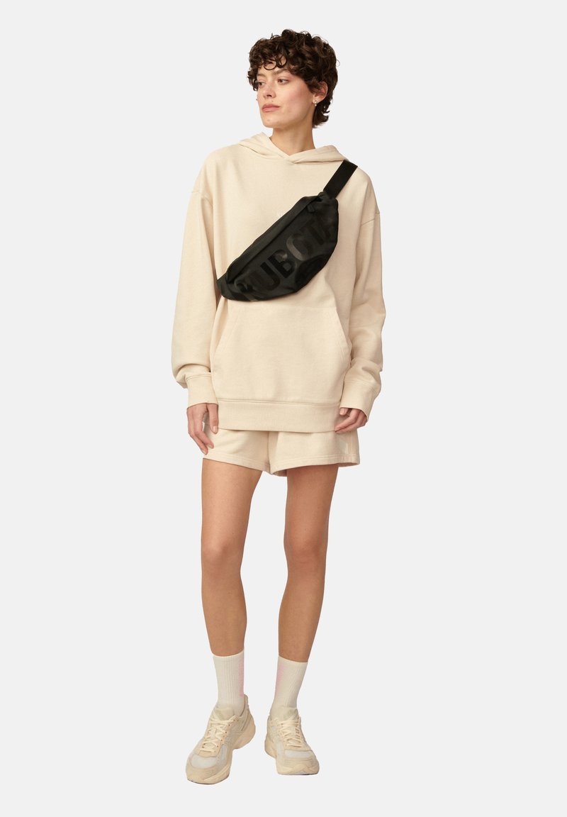 Beżowa, oversize'owa bluza z kapturem z kieszenią z przodu, w połączeniu z beżowymi szortami. Czarna nerka z grafiką "SUB", białe skarpetki i beżowe sneakersy.