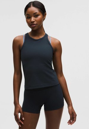 Ung kvinde iført sort, ærmeløs træningstop og sorte shorts, stående med afslappet kropsholdning mod en ensfarvet, lys baggrund.