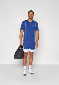 Tricou sportiv albastru cu accente turcoaz, pantaloni scurți albastre asortați cu dungi albastre deschise, ținând o geantă de voiaj neagră, purtând adidași albi.