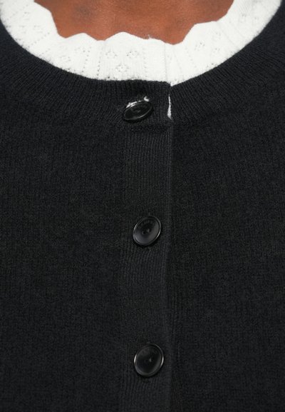 Černý pletený cardigan s čtyřmi černými knoflíky a žebrovaným límcem, přes bílou texturovanou blůzu s vlnitými okraji.