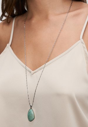 Silberne Halskette mit einem ovalen grünen Steinanhänger, strukturiert mit einer glatten Oberfläche, getragen über einem hellfarbenen Oberteil.