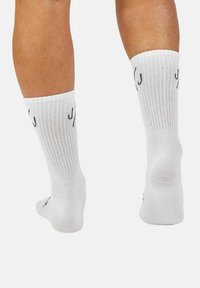 Weiße Socken mit gerippter Textur und schwarzem Schriftzug an der Seite. Entworfen für Komfort, zeigen sie den Fersen- und Knöchelbereich.