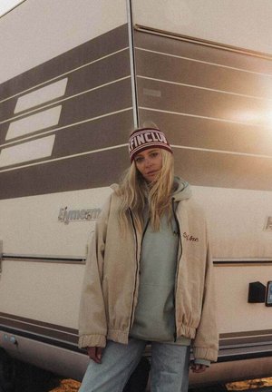 Młoda kobieta w casualowym zimowym ubraniu stoi oparta o beżowo-brązowy pasiastego kampera o zachodzie słońca.