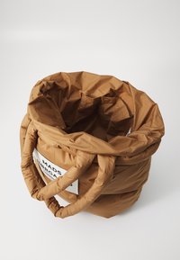 Barna, párnázott textil táska henger alakban, két fogantyúval és egy logó címkével az egyik oldalán. Észlelhető textúrált felületi részletek.
