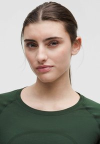 Młoda kobieta o jasnej karnacji i brązowych włosach związanych z tyłu, ubrana w ciemnozielony t-shirt z okrągłym dekoltem, patrzy bezpośrednio w obiektyw z neutralnym wyrazem twarzy.