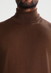 Brun polotröja i mjukt, elastiskt tyg med en figurnära design och hög krage. Texturen är ribbad och mjuk.