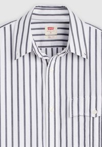 Camisa branca de botão com riscas verticais cinza escuro. Apresenta gola aberta, bolso frontal e botões brancos. A textura do tecido parece suave.