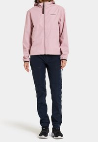 Personne portant une veste à capuche rose clair, un pantalon cargo bleu marine avec des poches zippées et des baskets noires, se tenant sur un fond blanc.