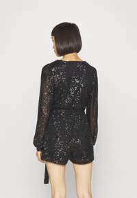 Svart paljett-romper med långa ärmar, som har ett bälte i midjan och en shortsdesign. Bakifrån visar den en slät, texturerad yta.
