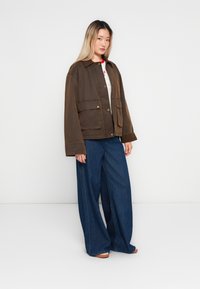 Jonge vrouw in een bruine jas, witte top en wijde blauwe jeans, staande tegen een effen lichtgrijze achtergrond.
