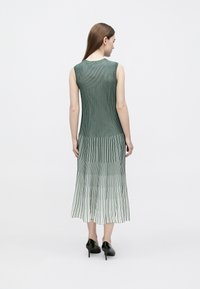 Vestido verde plissado sem mangas com riscas verticais, que passam para um tom mais claro na bainha. Textura suave, comprimento até meio da perna, usado com saltos pretos.