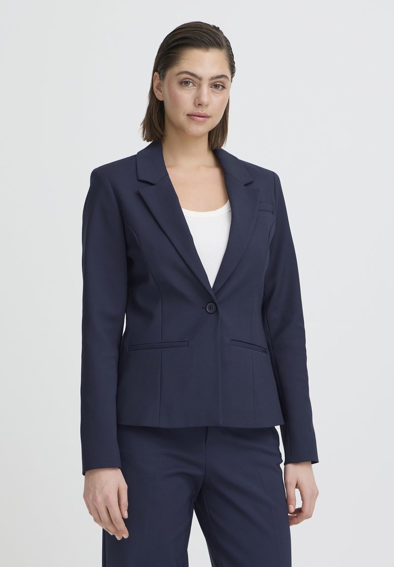 Femme debout portant un blazer ajusté bleu marine sur un haut blanc et un pantalon bleu marine assorti, devant un fond clair uni.