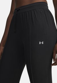 Zwarte sportieve broek met een koord in de taille, een gladde textuur, zijzakken en een zilveren logo aan de linkerkant van de voorkant.